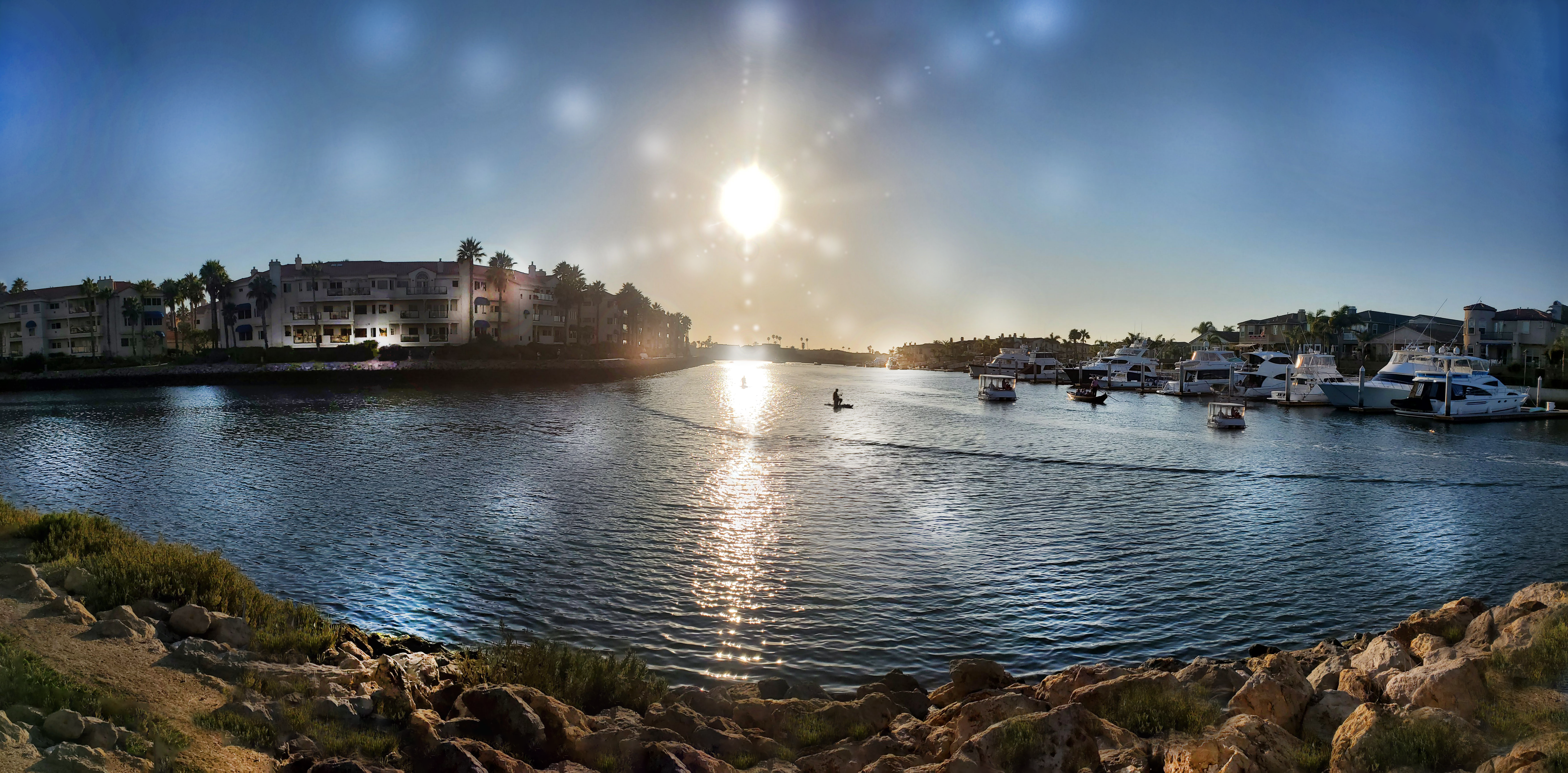 Panorama of a pier with hotels and palm trees on left side and boats parked on the right side, rocks in the front, sun beaming the center of the sky.