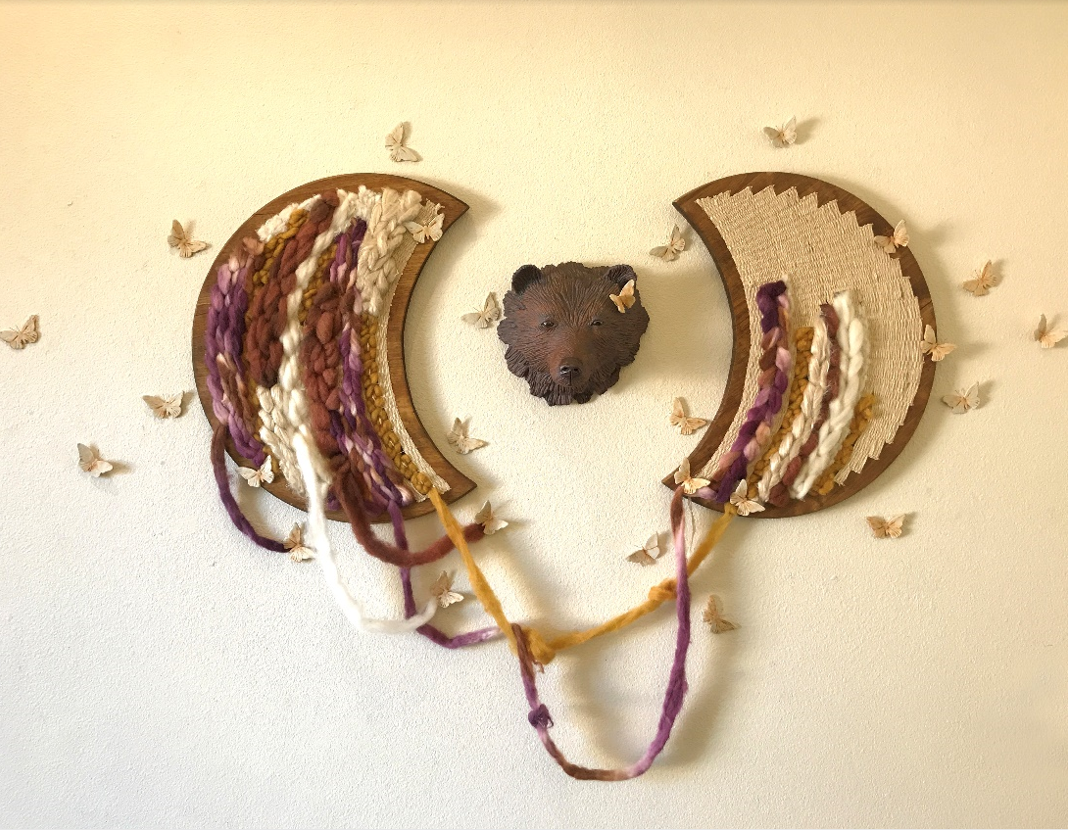 Ceramic bear head in the center with two wooden half moon shapes on each adjacent side with butterflies scattered around and fiber shaped as ropes hanging from the wooden pieces