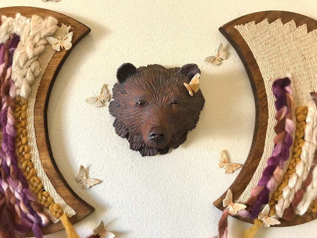 Close up of ceramic bear head in the center with two wooden half moon shapes on each adjacent side with butterflies scattered around and fiber shaped as ropes hanging from the wooden pieces